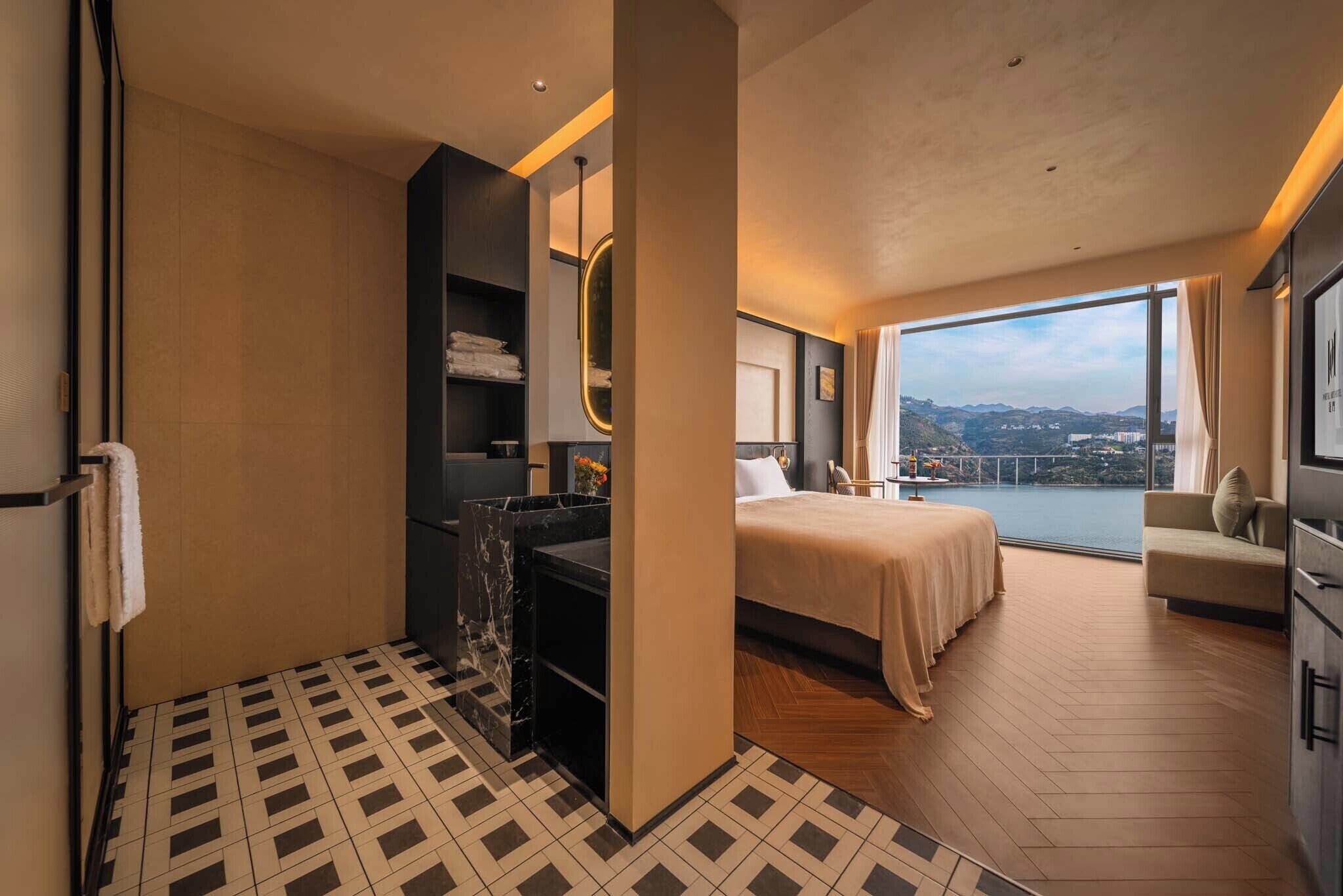 Una habitación de hotel moderna con una cama grande, un área de descanso con vistas, un baño con tocador de mármol y un amplio ventanal con vista a un paisaje urbano.