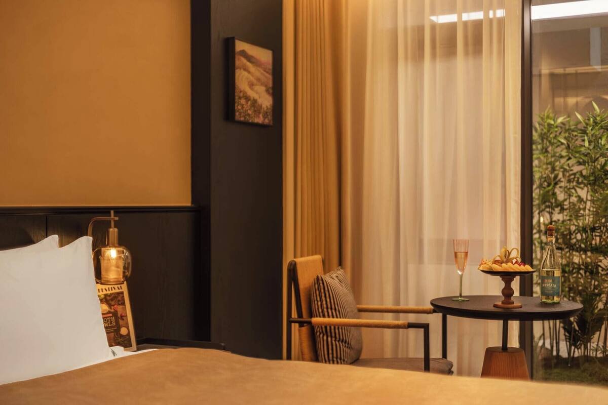 Une chambre d’hôtel avec un lit, une chaise, une petite table avec un verre et une bouteille, et une fenêtre avec des rideaux.