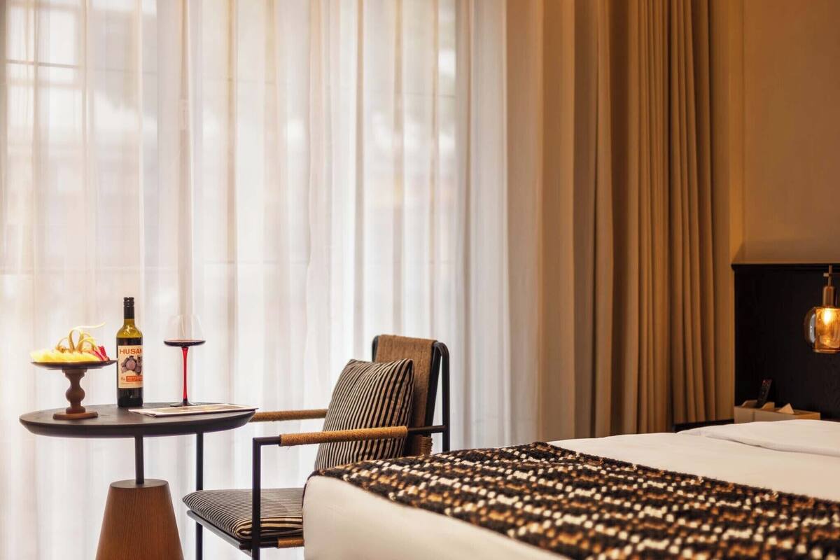 Une chambre d’hôtel avec un lit, une chaise, une petite table sur laquelle se trouvent une bouteille et un verre, et des rideaux légers.