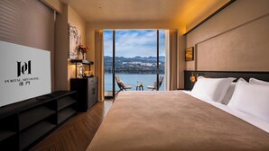 Une chambre d’hôtel avec un grand lit, un bureau, une chaise, une petite table et une vue sur un plan d’eau et des montagnes.