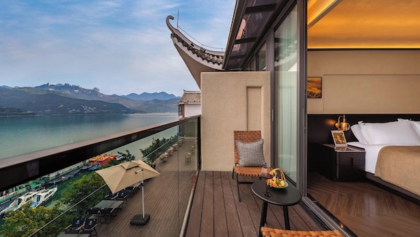 Un balcon avec vue sur un lac et des montagnes, un lit avec une table de chevet, et une petite table avec une lampe.