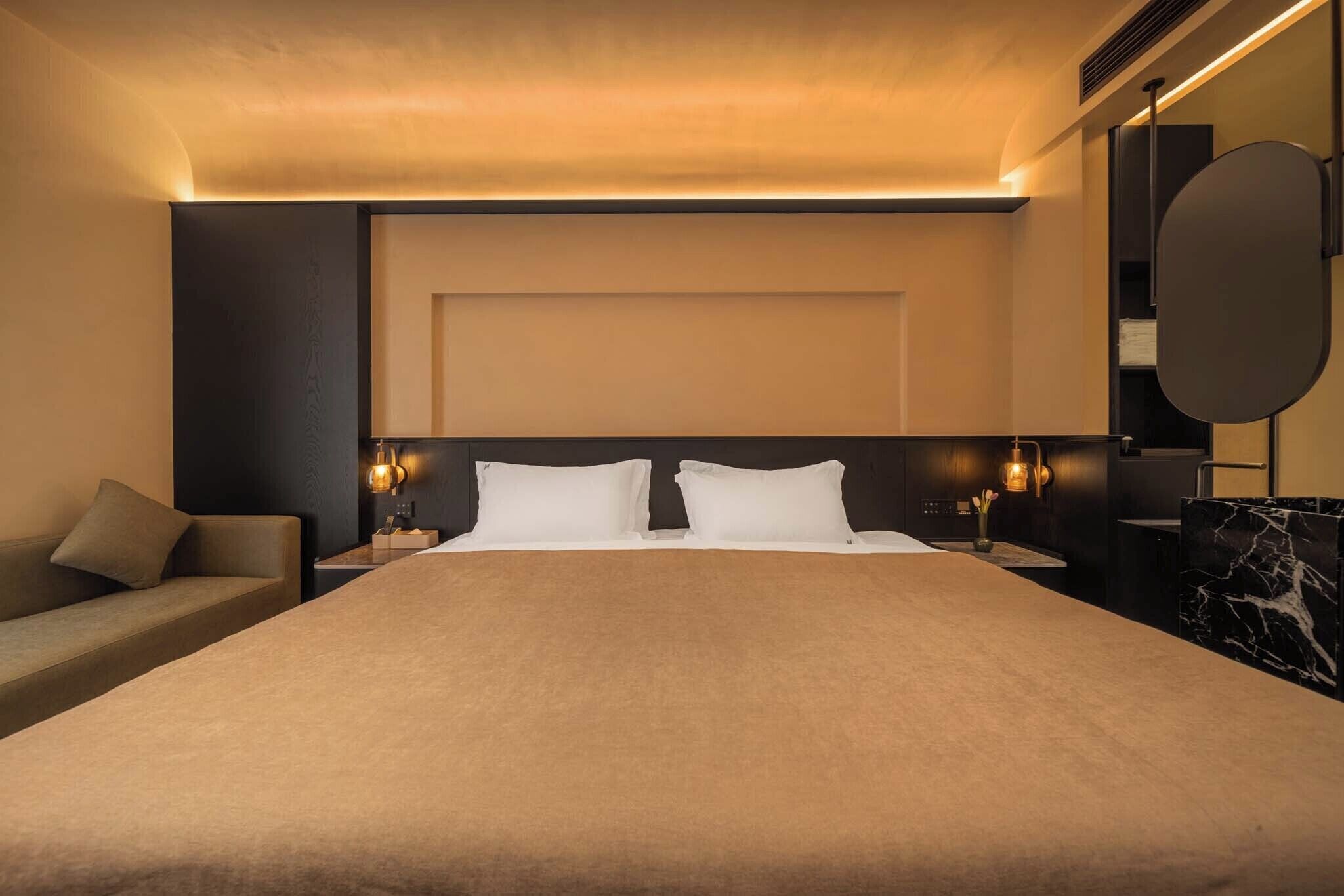 A modern hotel room with a large bed, a sofa, a nightstand with a lamp, and a mirror.