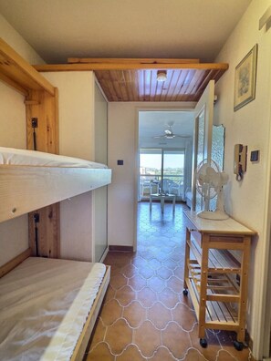 Een kamer met een stapelbed, een ventilator, een klein tafeltje en uitzicht op een balkon met een plafondventilator.