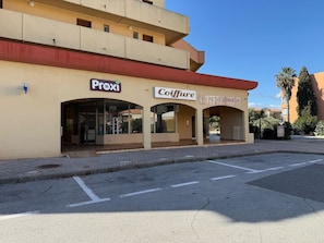 A building with 'Proxi' and 'Coiffure' signs, a covered entrance, and a clear sky. - One-bedroom apartment with a sleeping nook, air conditioning, and a view of the sea (La Londe-les-Maures)