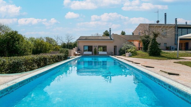 Una piscina con superficie d'acqua cristallina e blu, circondata da un giardino curato e un edificio con tetto in tegole.