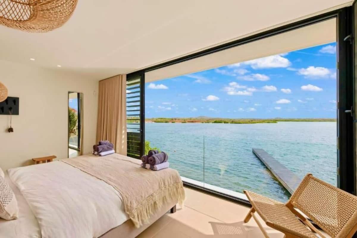 Camera da letto con una grande finestra vista su un corpo d'acqua, un letto, una sedia e una lampada in vimini.