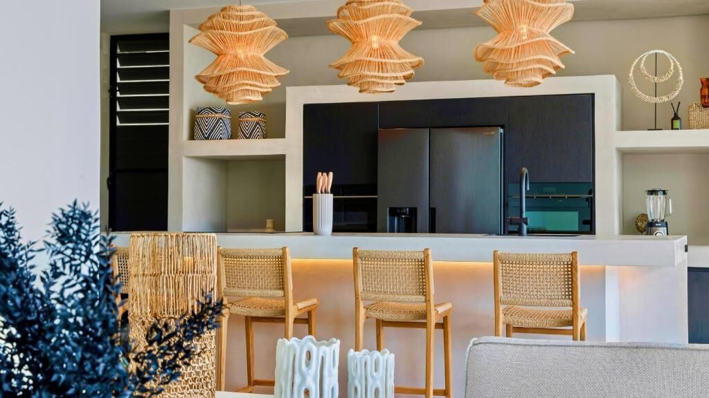 A modern kitchen with a black refrigerator, a built-in oven, and a coffee machine. There are three wicker chairs and a white sofa in the living area.