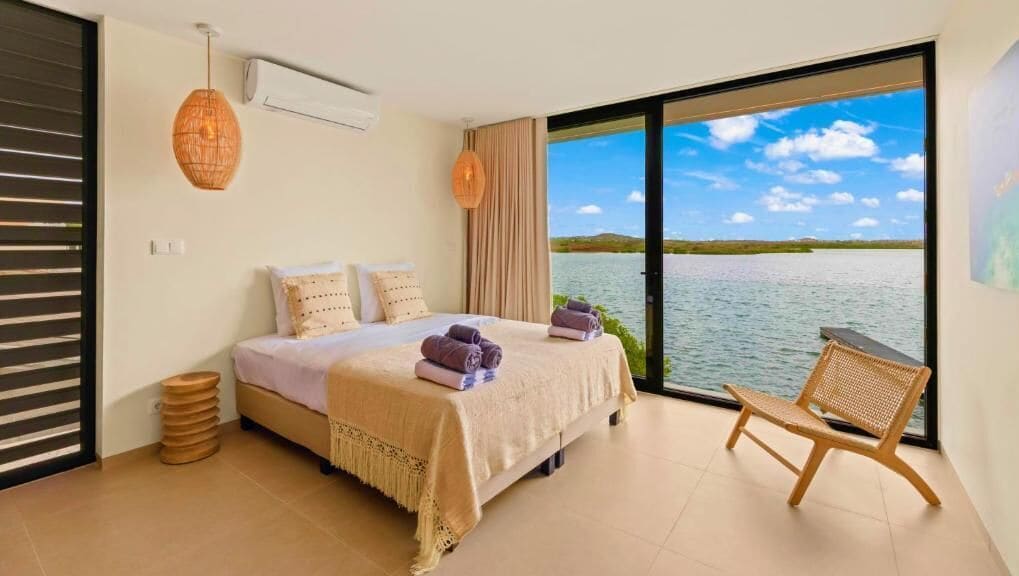 A bedroom with a bed, a chair, a view of water, and a large window.