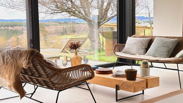 A room with a large window, a wicker chair, a sofa, and a wooden coffee table. - Luxury Villa with Garden & Zipline (La Roche-en-Ardenne)