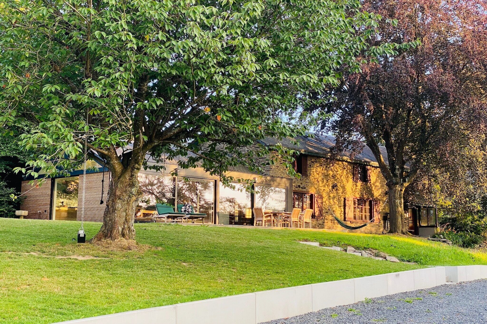 Una casa con un árbol grande en el jardín delantero, un patio con mobiliario exterior y un edificio de piedra al fondo.