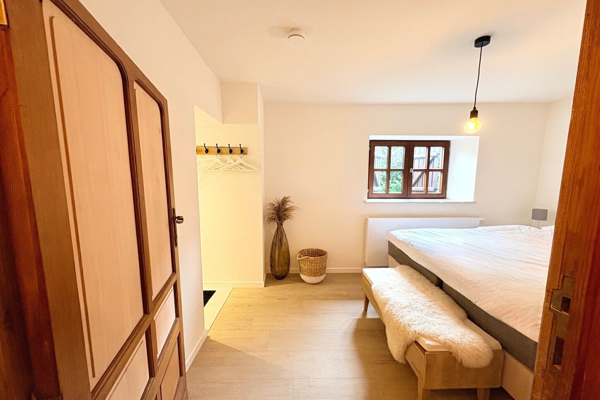 A bedroom with a bed, a bench, a window, a hanging light, and a door.