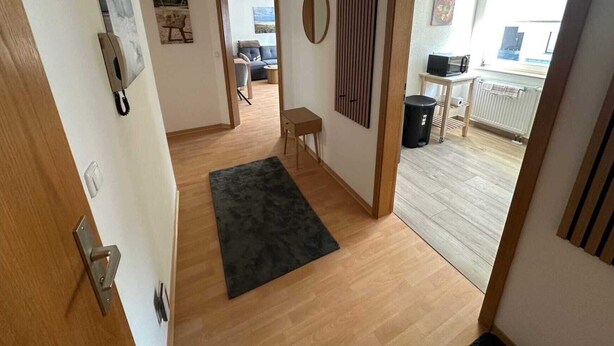 A hallway with a wooden floor, a black rug, a door with a silver handle, and a wall with a circular decoration. - behagliche Ferienwohnung in Elstra (Elstra)