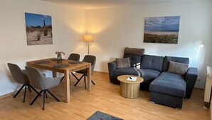 A modern living room with a wooden dining table, grey upholstered chairs, a grey sofa, a small round coffee table, and a grey rug. - behagliche Ferienwohnung in Elstra (Elstra)
