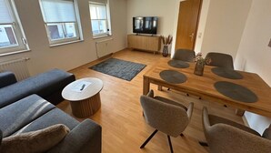 A modern living room with a sofa, a coffee table, a dining table with chairs, a TV, and a radiator. - behagliche Ferienwohnung in Elstra (Elstra)