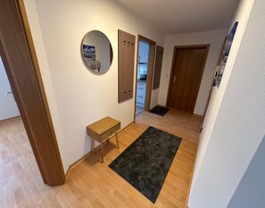 Ein Korridor mit Holzboden, einem kleinen Holztisch, einem schwarzen Teppich, einem runden Spiegel und einer Holztür.
