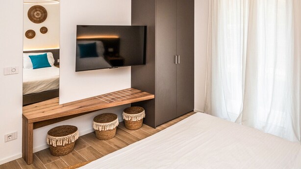 A bedroom with a bed, a TV mounted on the wall, a wardrobe, and a bedside table with baskets. - Pop Vibes - Ariom Apartments (Moniga del Garda)
