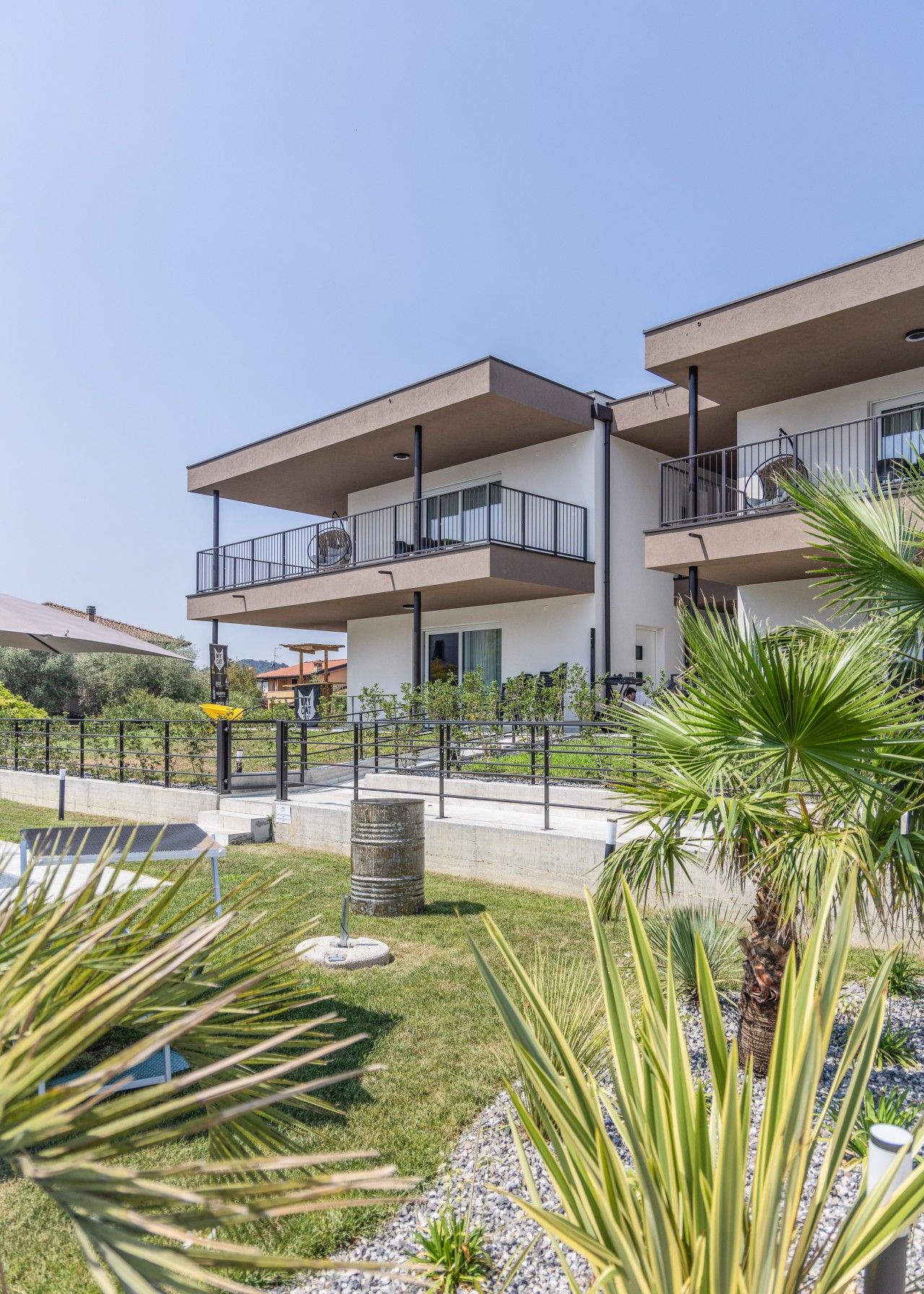 A modern multi-story building with balconies, a paved courtyard, and surrounding greenery.