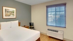 Une chambre d’hôtel avec un lit, un tableau encadré au mur, une fenêtre avec des stores et un climatiseur fixé au mur.