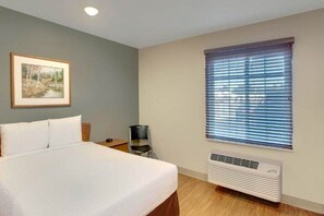 A hotel room with a bed, a framed picture on the wall, a window with blinds, and a wall-mounted air conditioner.