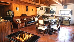 A cozy living room with a wooden ceiling, a brown leather sofa, a wooden coffee table with a chess set, and a dining area with chairs and a table.