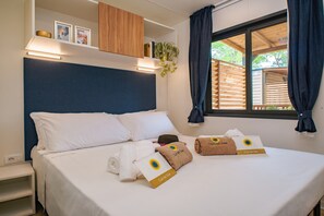 Une chambre d’hôtel avec un lit, une table de chevet, une étagère avec des photos et une plante, et une fenêtre avec des rideaux.