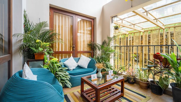 A cozy indoor seating area with a sofa, chairs, and a coffee table, surrounded by potted plants and a glass door leading to a balcony. - Alara by Alaya Stays 4-BHK Apartment (Kanayannur)