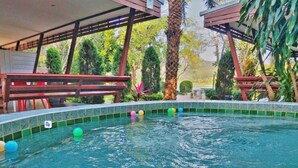 Una piscina con pelotas de colores flotando, rodeada por una zona cubierta con vigas de madera y follaje.