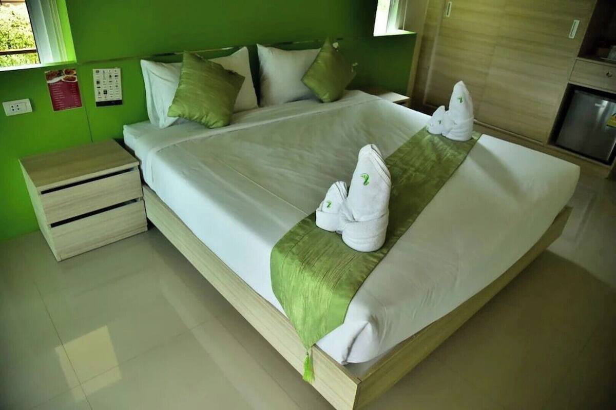 Una cama bien hecha con sábanas blancas y almohadas verdes, una mesita de noche de madera y una pequeña mesa con un menú en la pared.