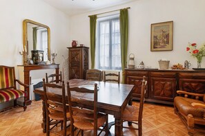 A dining room with a wooden table and chairs, a mirror, a radiator, and a window with green curtains. - Apartment “Hortensia” with Private Terrace and Garden (Sarlat-la-Canéda)