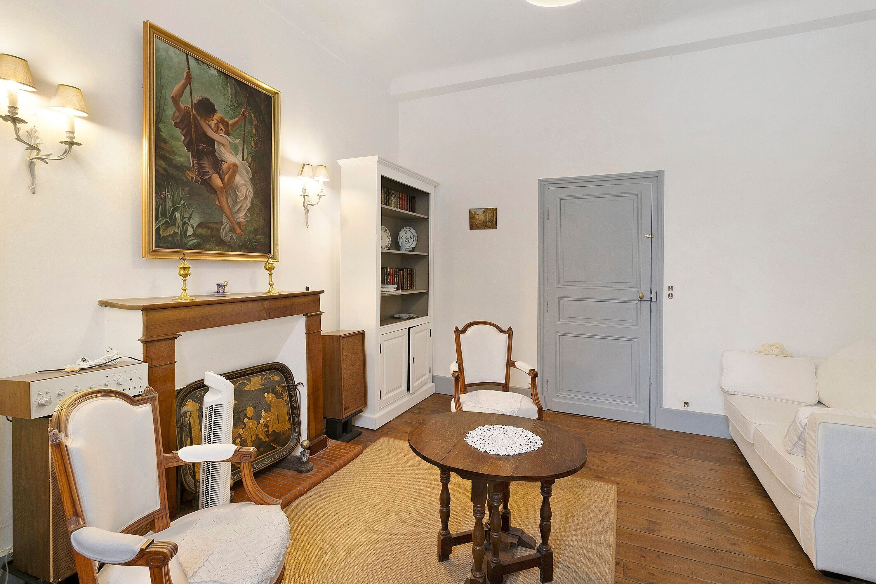 A room with a round wooden table, two white upholstered chairs, a white sofa, a fireplace with a mantel, a bookshelf, and a door.