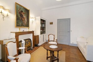 A room with a round wooden table, two white upholstered chairs, a white sofa, a fireplace with a mantel, a bookshelf, and a door. - Apartment “Hortensia” with Private Terrace and Garden (Sarlat-la-Canéda)