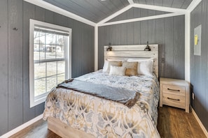 A bedroom with a wooden headboard, a bed with patterned bedding, a nightstand, and a window with a view. - Gilbertsville Hideaway: 1 Mi to Lake! (Gilbertsville)