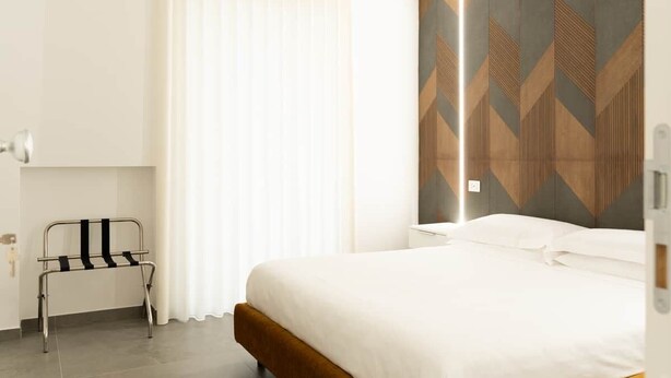 A hotel room with a bed, a bedside table, a chair, and a window with curtains. - Flamingo affittacamere - Camera 4 (Matera)