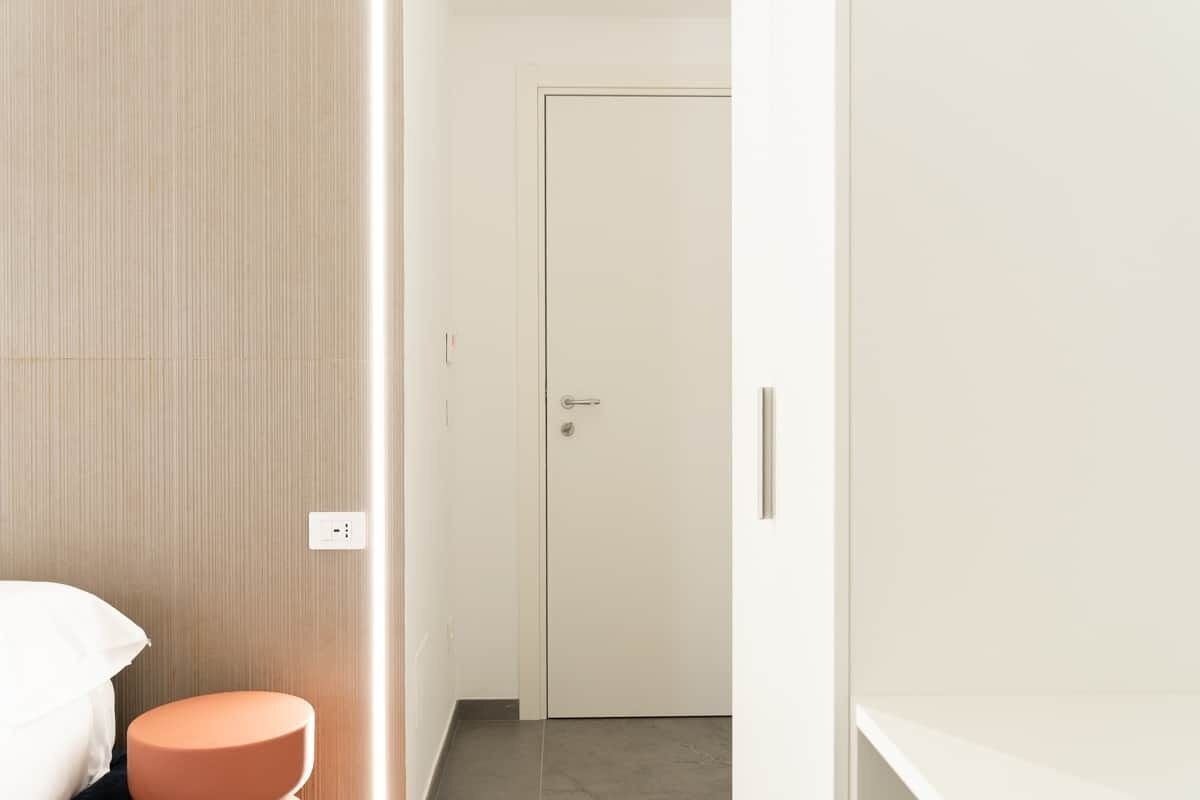 A hotel room with a bed, a small orange stool, a white door, and a light switch on the wall.