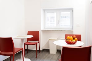 Un coin repas avec une table ronde, deux chaises rouges, une fenêtre avec des stores et un saladier de fruits posé sur la table.