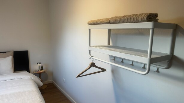 Une chambre avec un lit superposé, une table de chevet avec une lampe et une étagère fixée au mur, sur laquelle se trouvent une serviette et un cintre.
