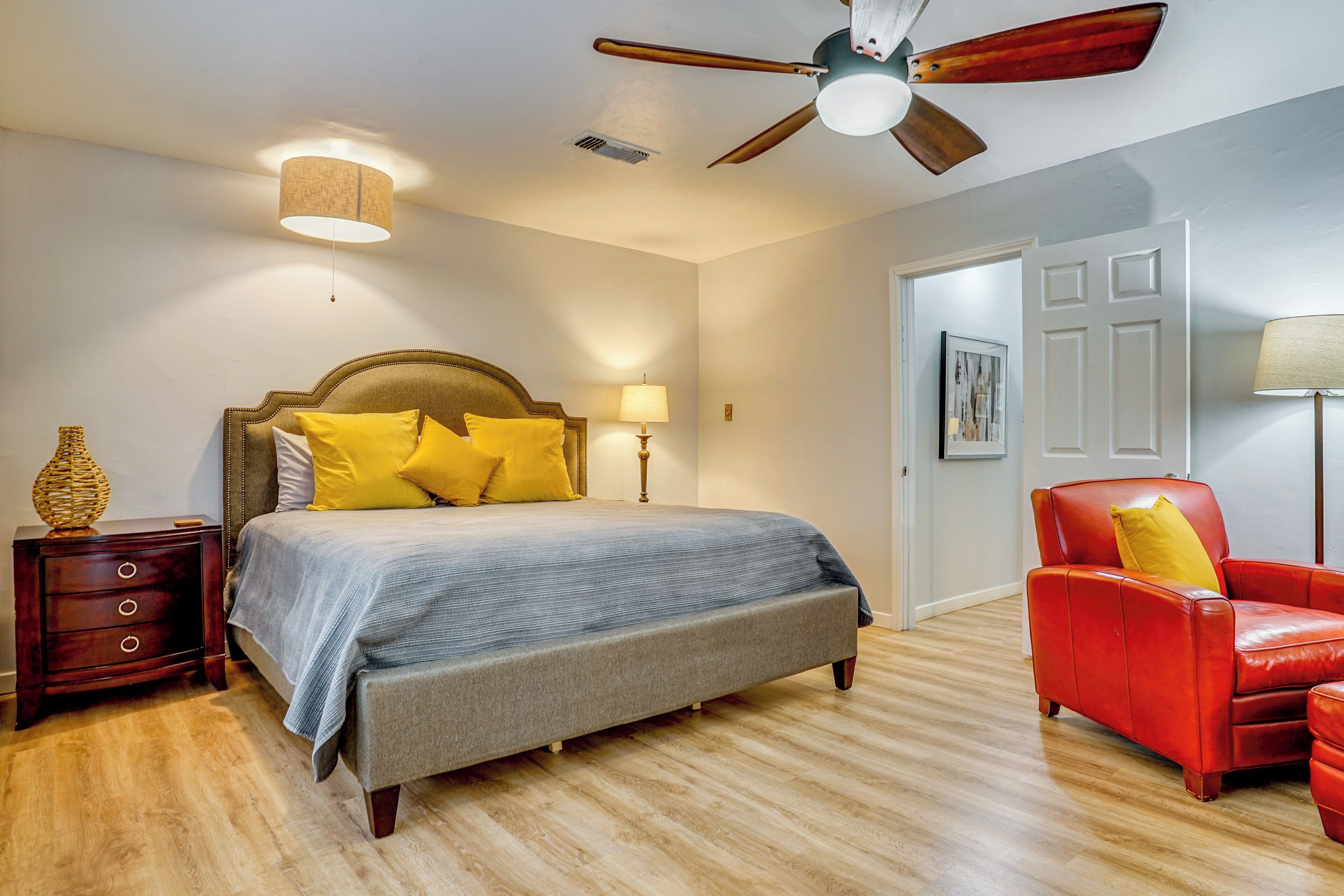 A bedroom with a bed, a nightstand, a red armchair, a ceiling fan, and a door.