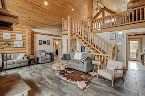 A log cabin interior with a staircase, wooden walls, a sofa, a chair, and a cowhide rug. - Group Retreat in the Bitterroot Valley w/ Hot Tub (Victor)