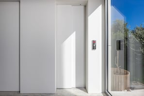A modern building entrance with a glass door, a small wooden planter, and a security device. - RM Grande Villa – Left Wing (Rovinj)