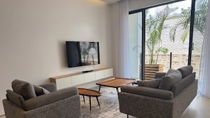 Een moderne woonkamer met twee grijze banken, een houten salontafel, een aan de muur bevestigde televisie en een groot raam met uitzicht op palmbomen.
