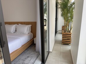 A bedroom with a bed, a wooden headboard, a grey carpet, and a view of a balcony with plants. - Omica 1 Residence (Kigali)
