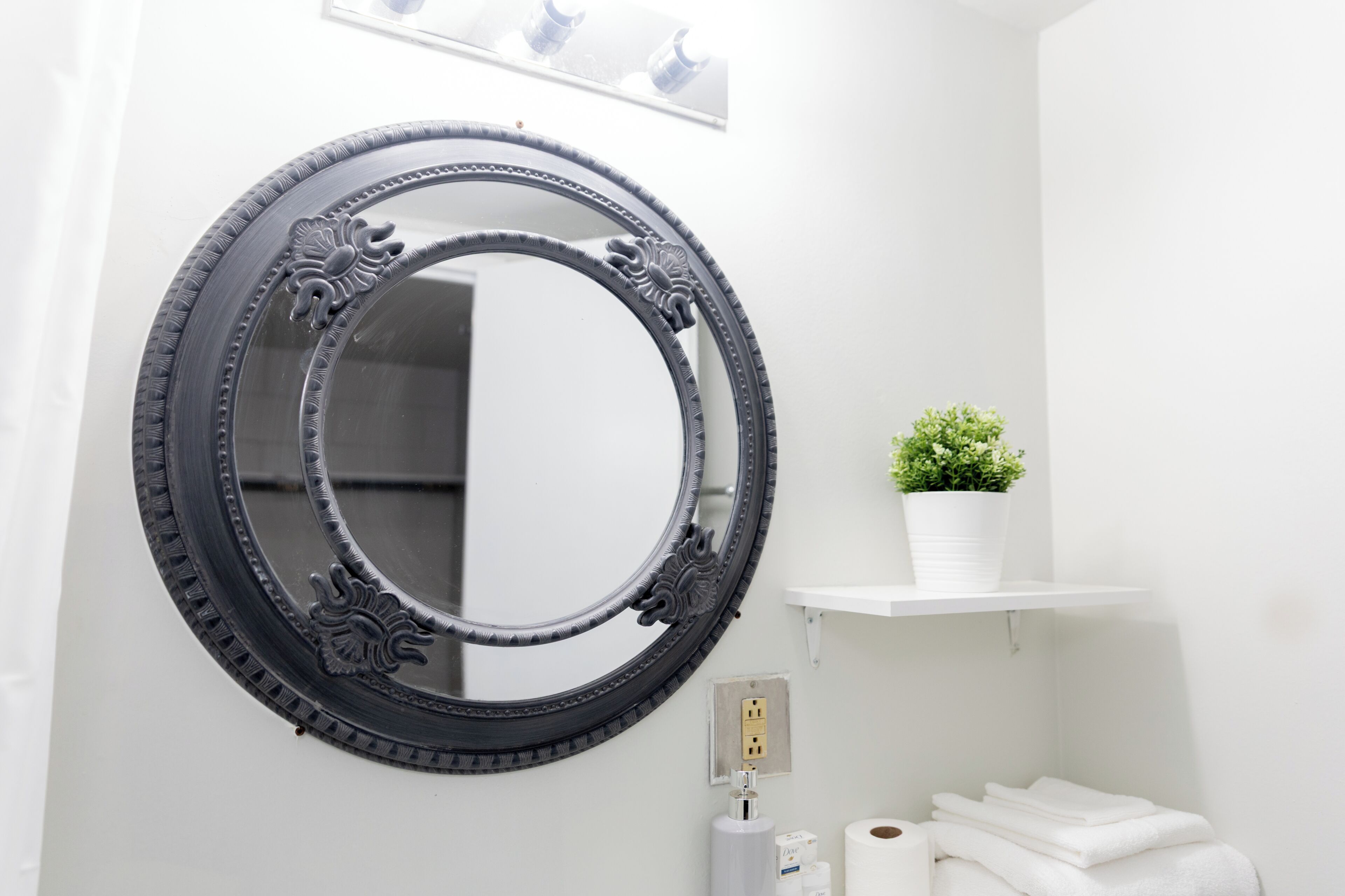 A bathroom with a round mirror, a potted plant, a white shelf, and a toilet paper holder.