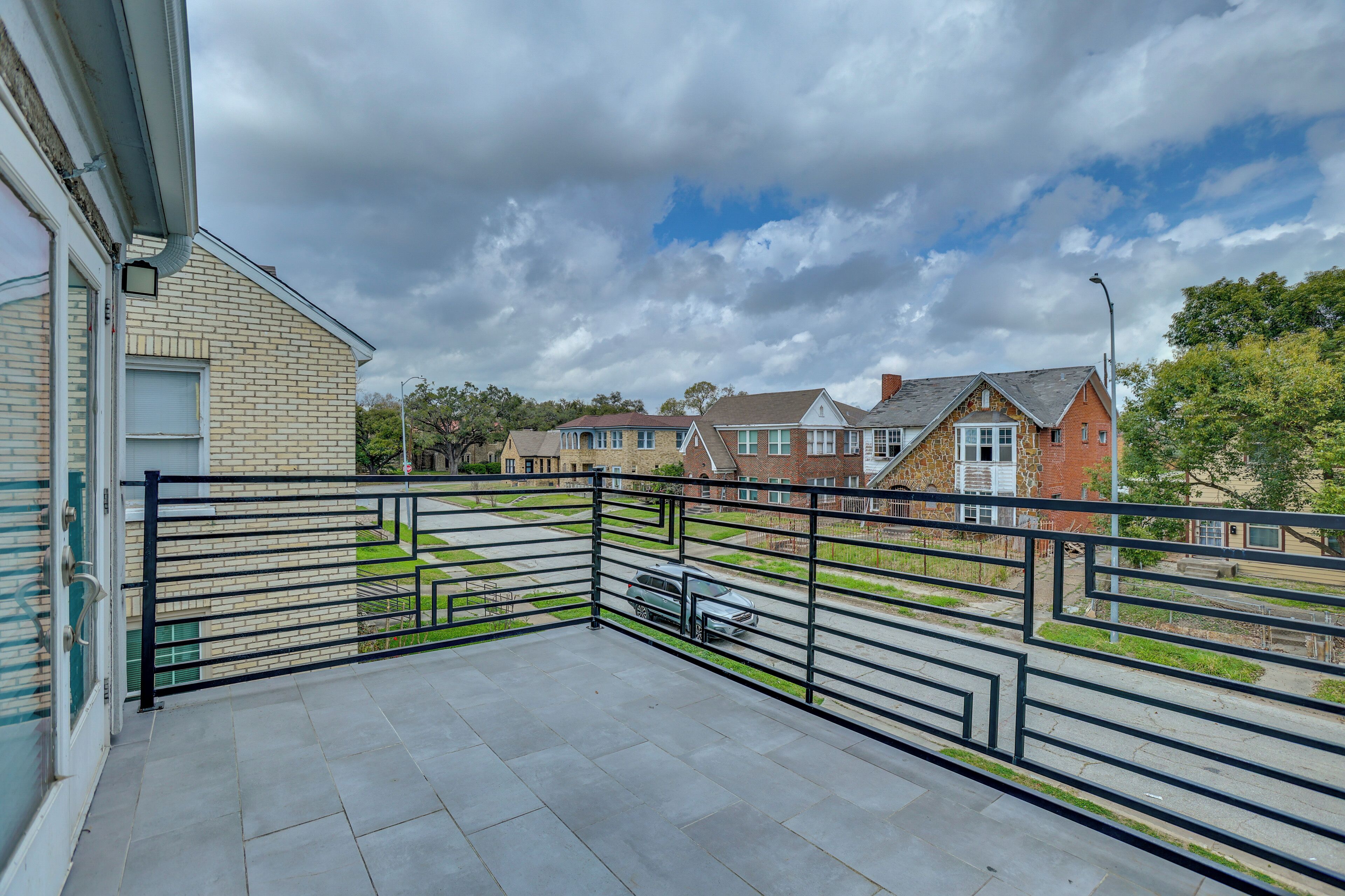 A balcony with a metal railing, a paved floor, and a view of residential buildings in the background.