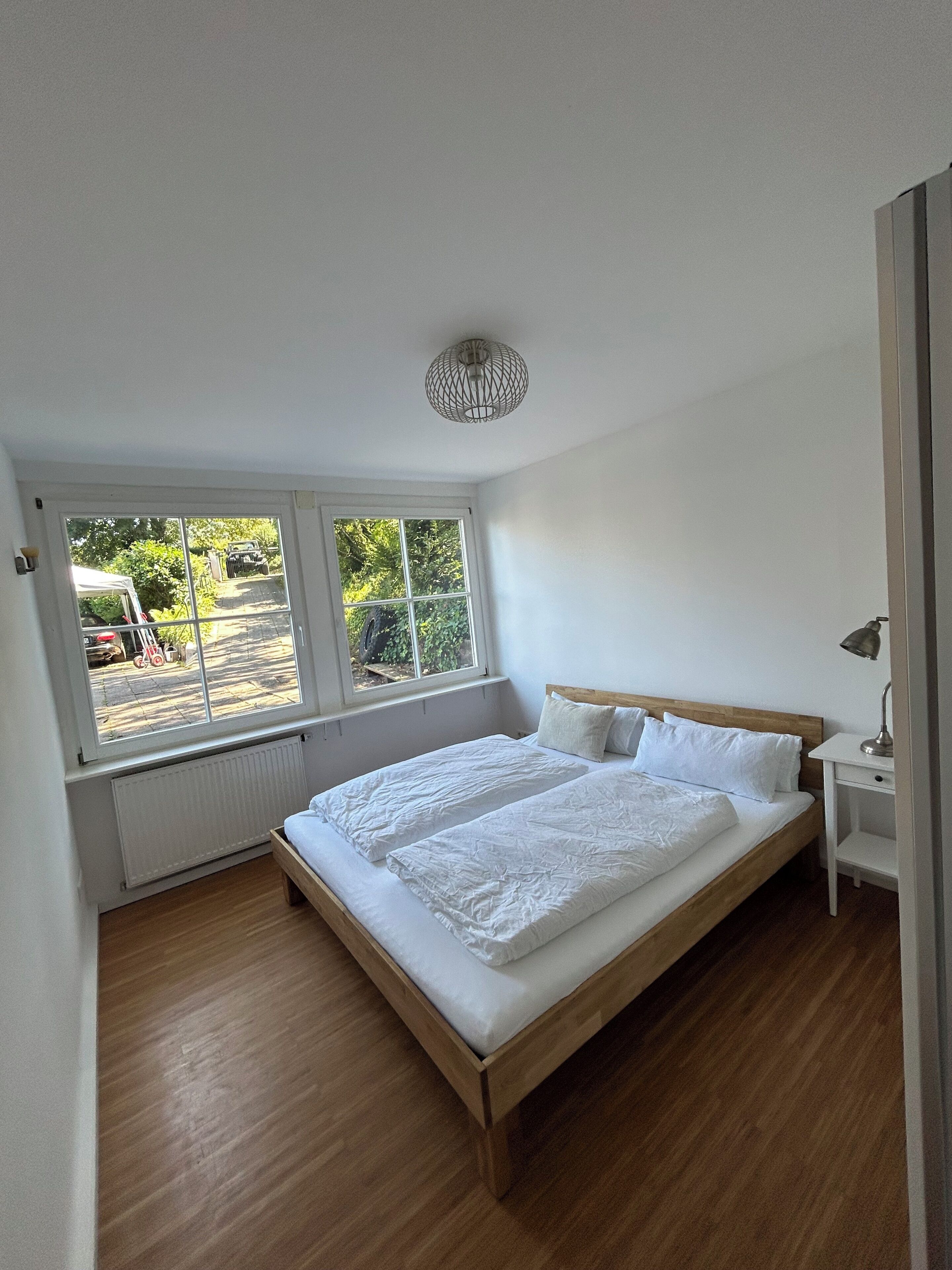 Un dormitorio con cama de madera, radiador blanco, ventana con vista a la calle y lámpara en el techo.