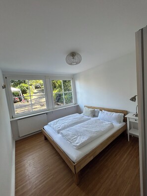 Une chambre avec un lit en bois, un radiateur blanc, une fenêtre donnant sur la rue et un luminaire au plafond.