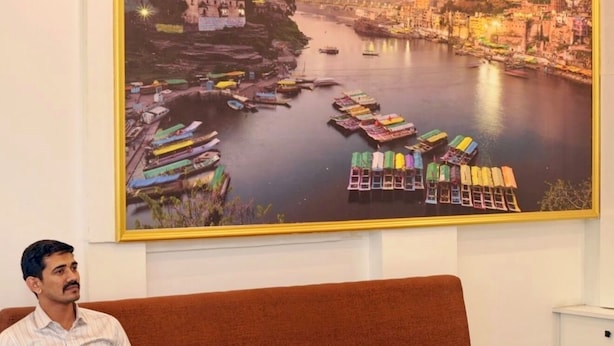 A man sitting on a brown sofa in a room with a large river scene painting on the wall. - The Mint Tree Hotels & Resorts (Bhopal)