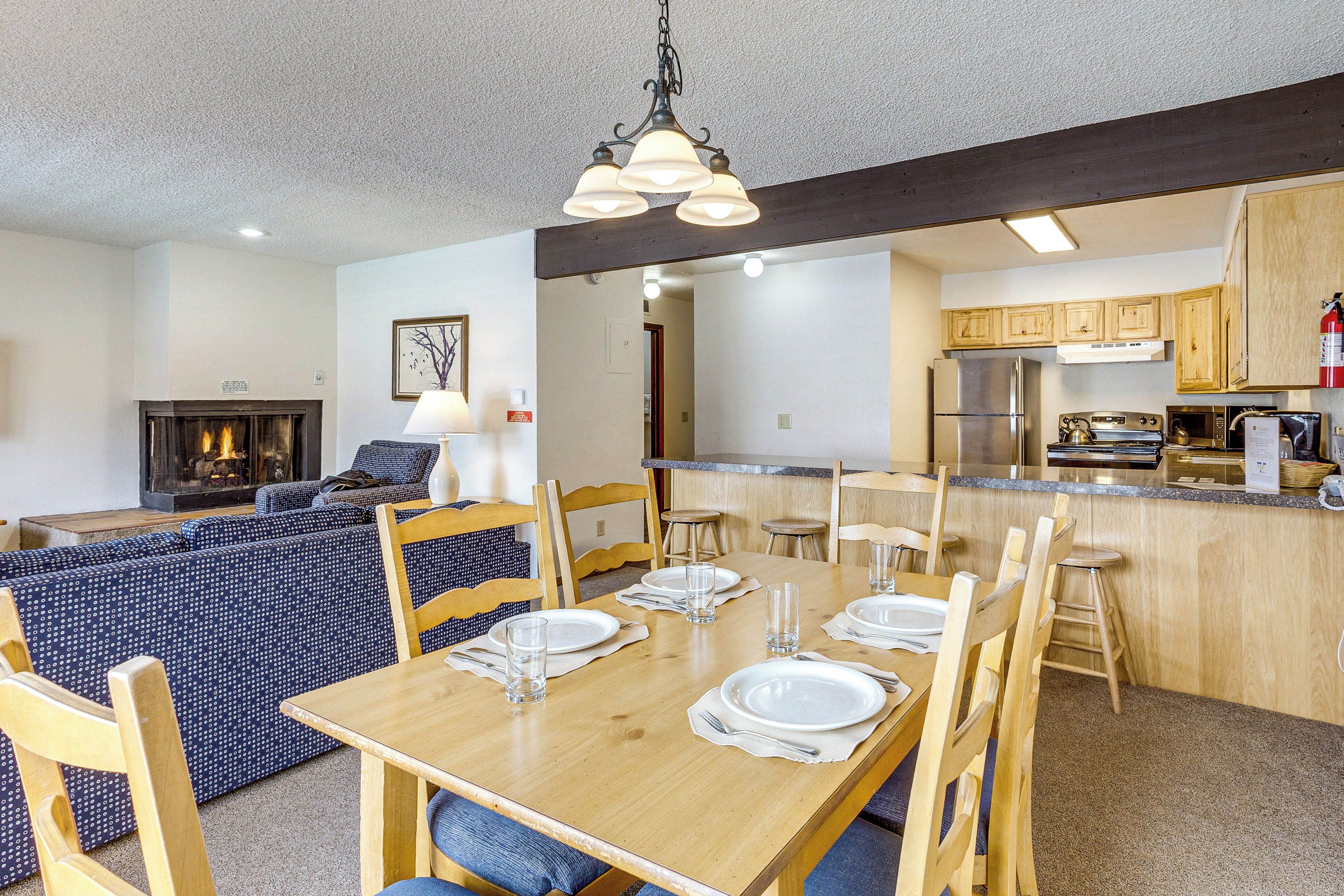 Een eetgedeelte met een houten tafel gedekt voor zes personen, een keuken op de achtergrond en een zithoek met een open haard.