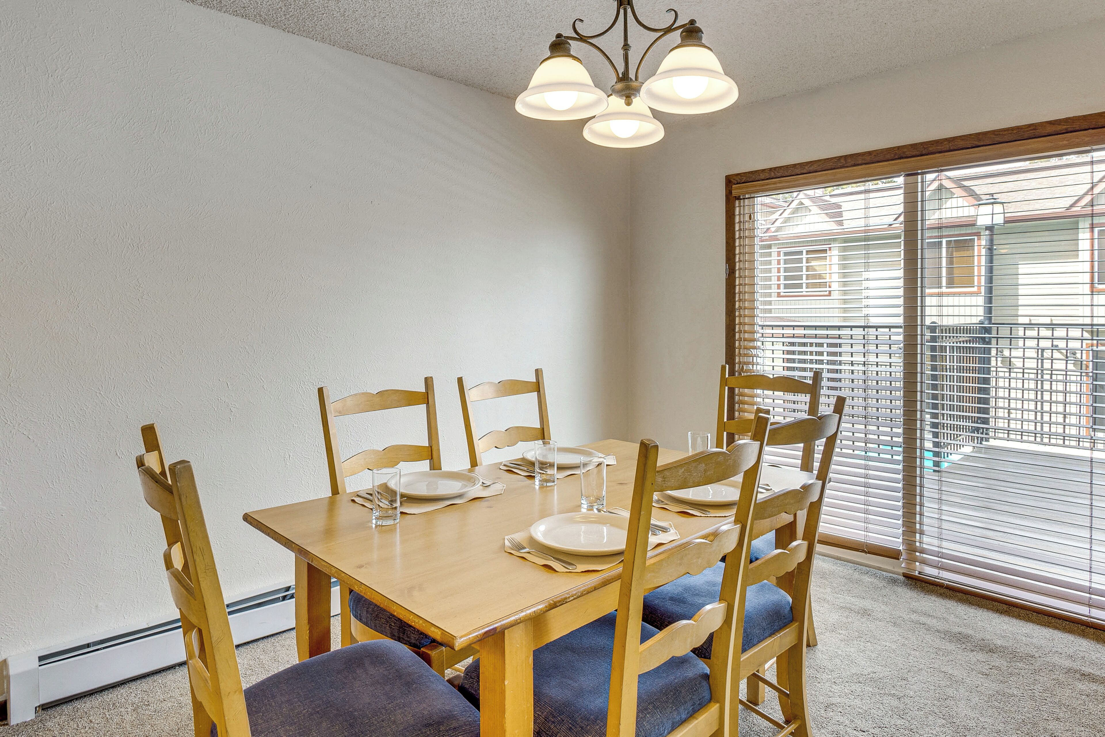 Comedor con mesa de madera dispuesta para cuatro, una araña y vistas a través de una ventana con persianas.