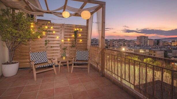 En træpergola med lyskæder, to stole med mønstrede hynder og en potteplante på en tagterrasse med udsigt over byen.