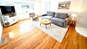 Ein Wohnzimmer mit grauem Sofa, einem Holztisch, einem Fernseher auf einem Schrank und einer weißen Ziegelwand.